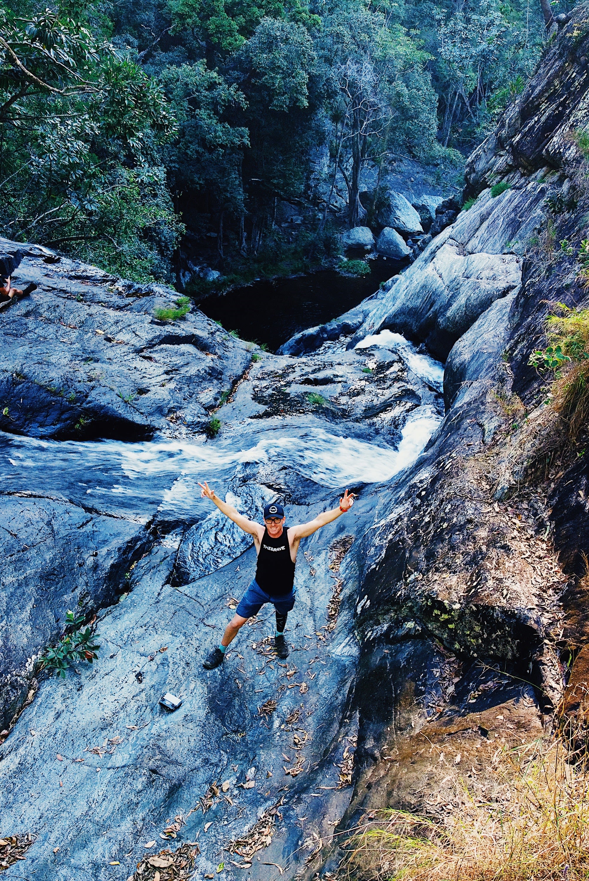 A man smiling and doing the peace sign. Behind him is a waterfall.